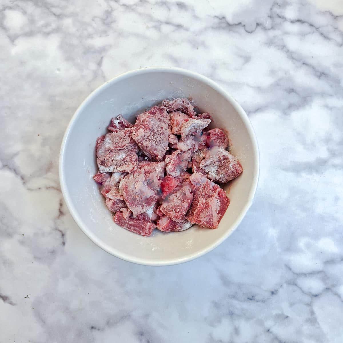 Pieces of beef coated with flour in a small white mixing bowl.