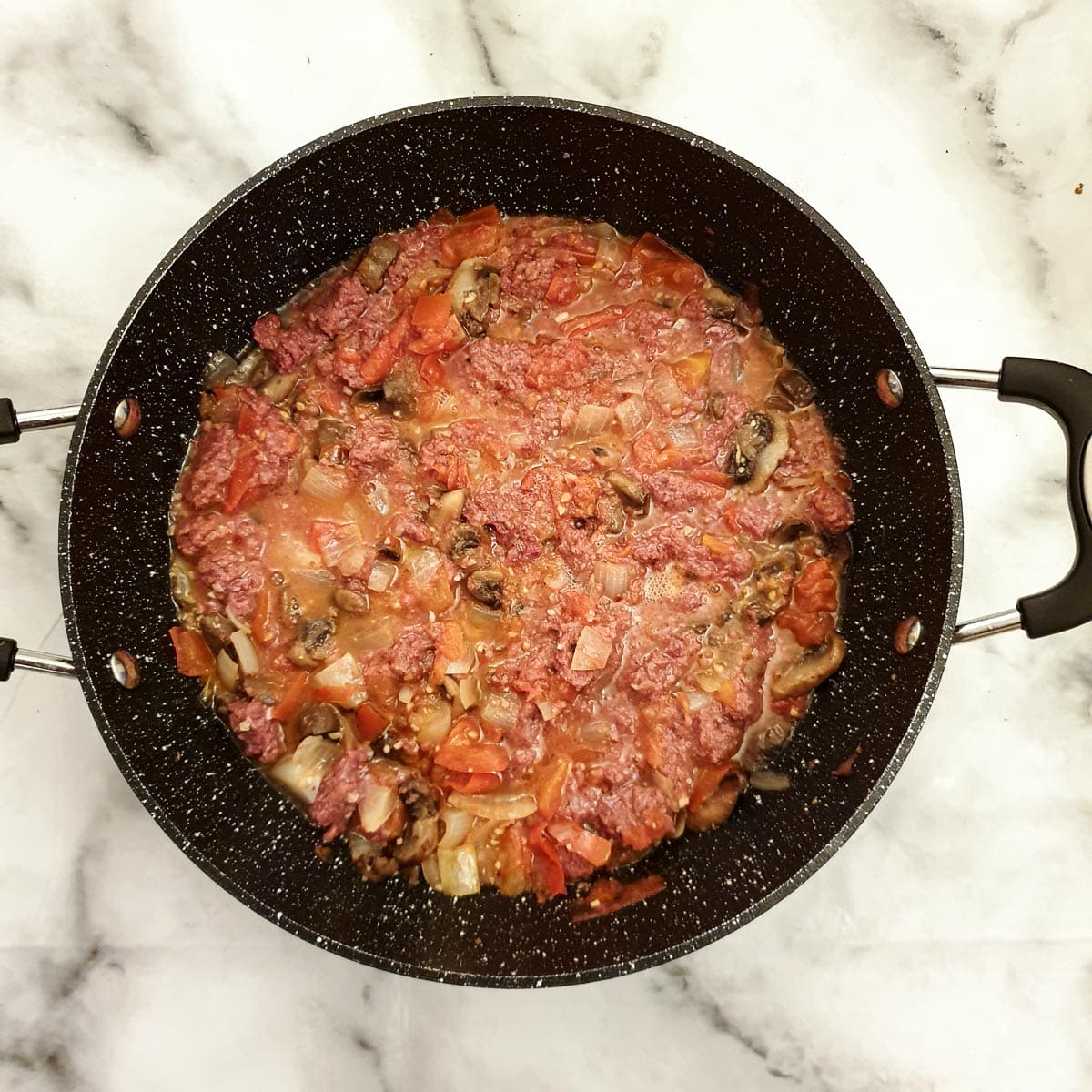 Bully beef stirred into tomatoes, onions and mushrooms in a frying pan.