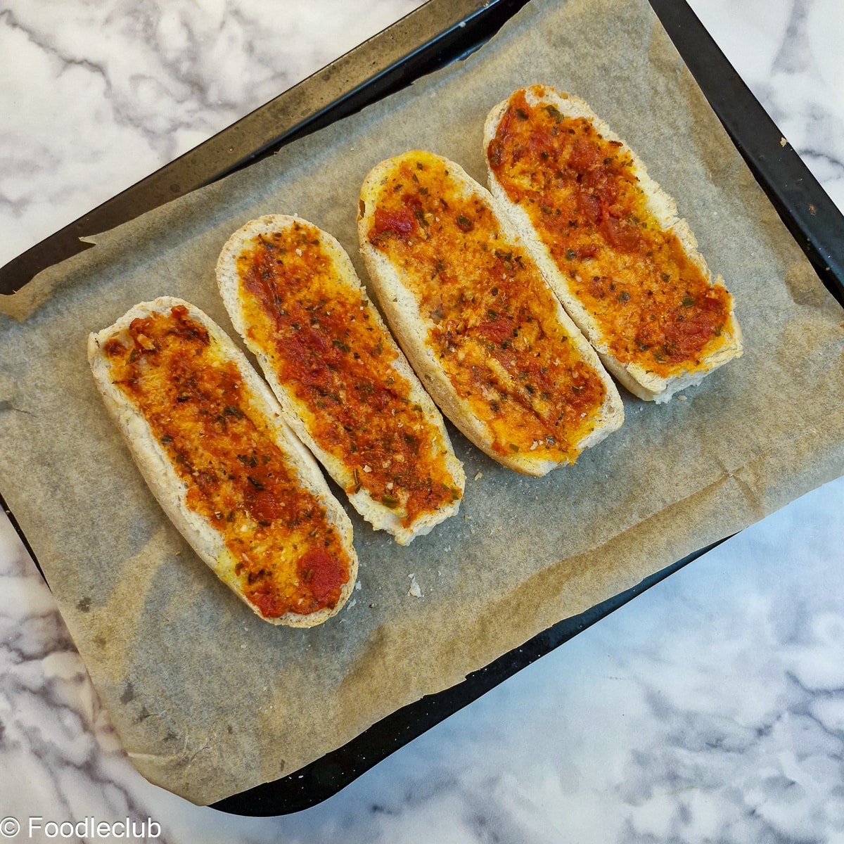 4 halved bread rolls spread with garlic butter on a baking tray after being spread with spaghetti sauce.