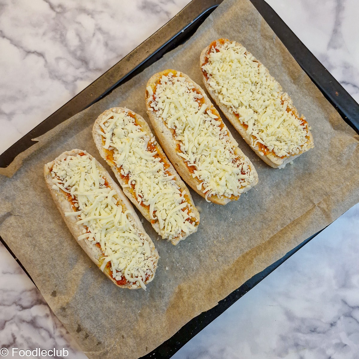 4 halved bread rolls spread with garlic butter on a baking tray topped with grated cheese.