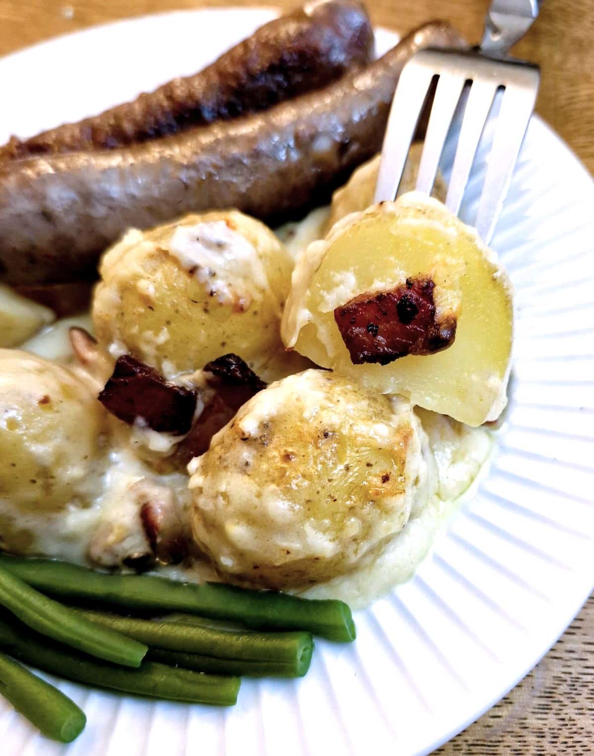 A helping of cheesy potato casserole on a plate with sausages and green beans.  One of the pieces of potato is held on a fork.