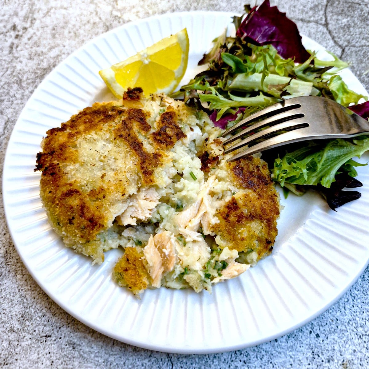 A salmon fishcake which has been cut open on a white plate with lettuce and lemon wedges.