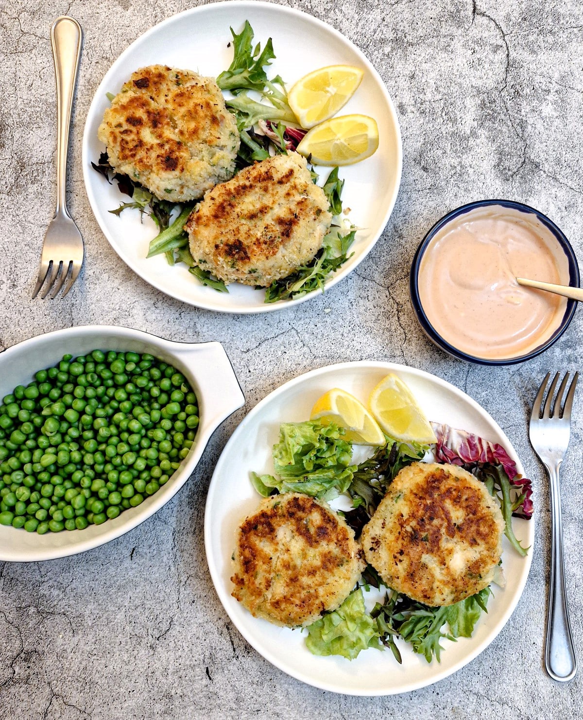 Overhead shot of two plates of salmon fishcakes with a dish of peas and a dish of seafood sauce.