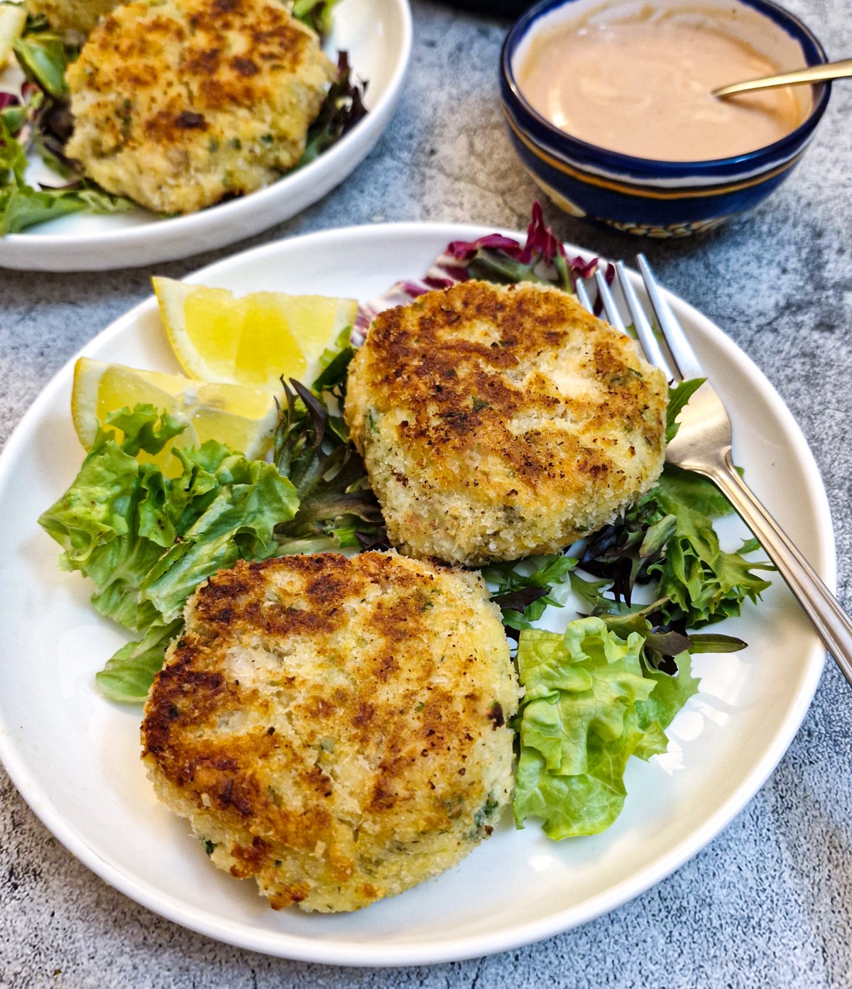 Two salmon fishcakes on a white plate on a bed of lettuce with lemon slices.