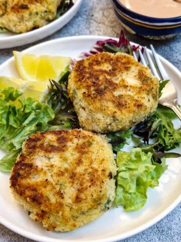 Two salmon fishcakes on a white plate on a bed of lettuce with lemon slices.