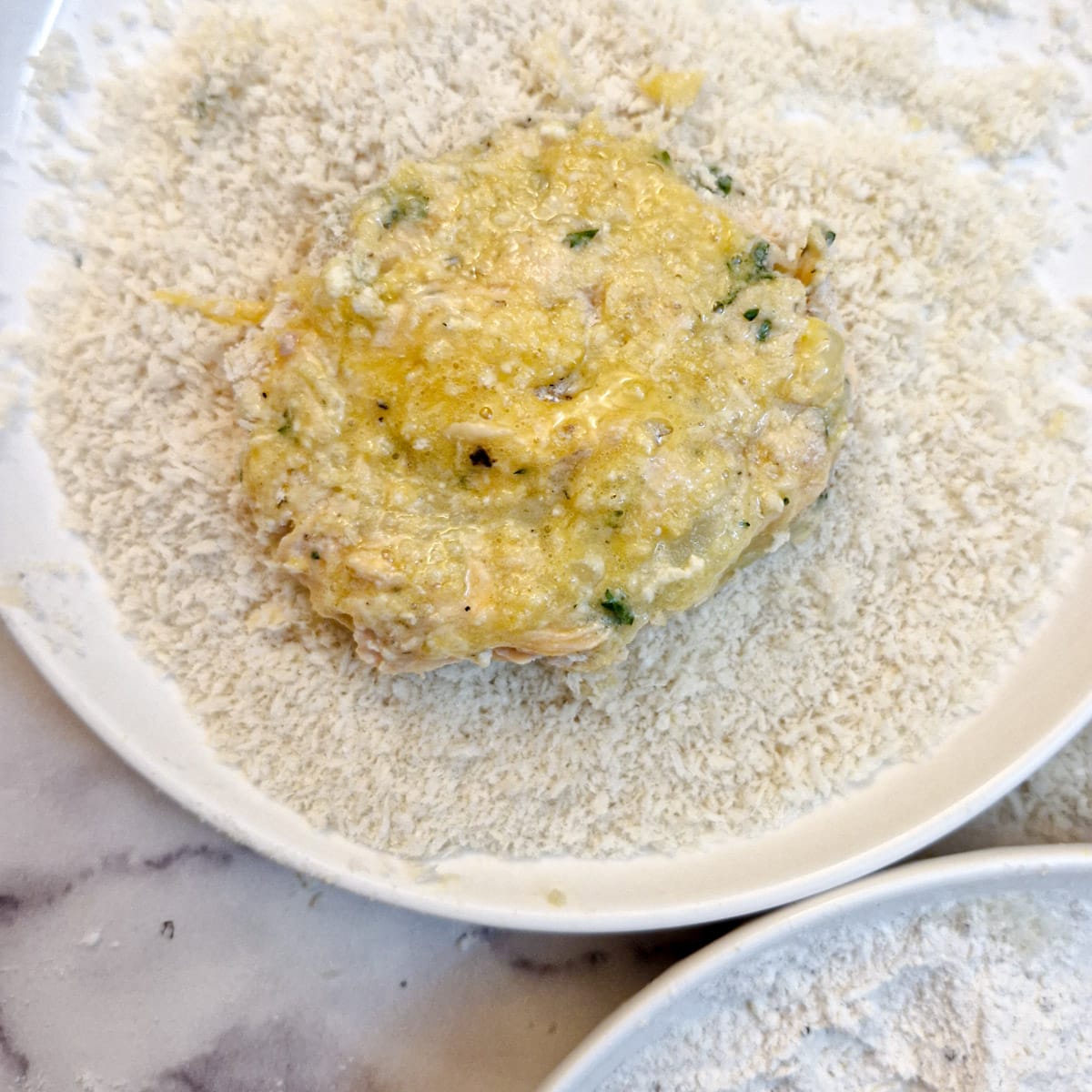 A fish patty in a bowl of breadcrumbs, brushed with beaten egg.