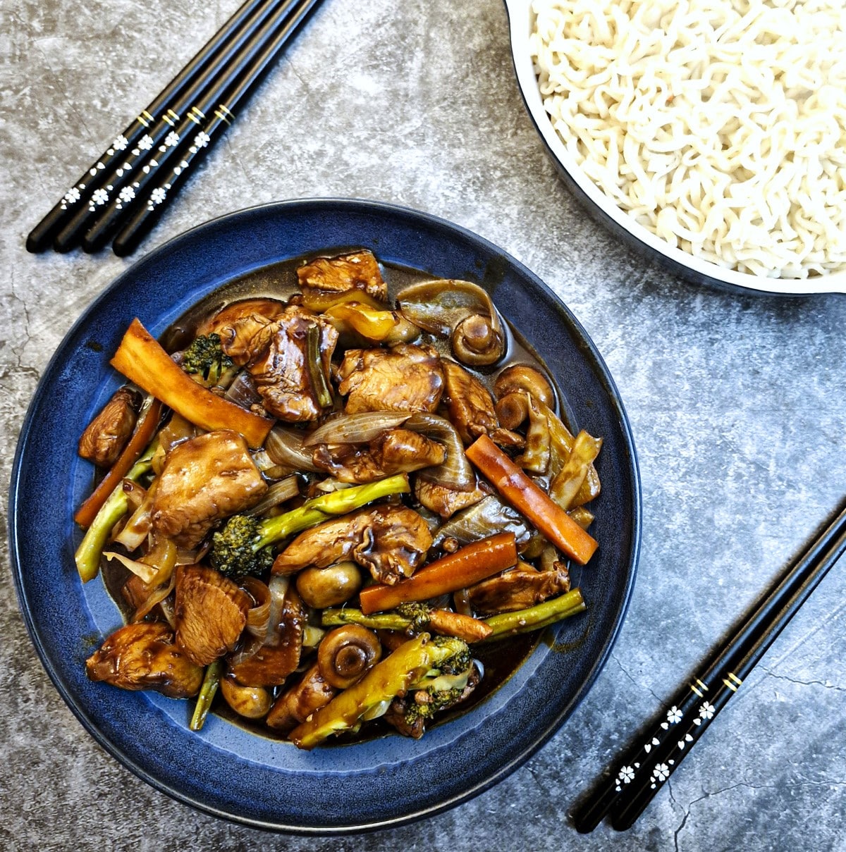 A blue serving dish of Szechuan chicken on a table next to a dish of noodles.