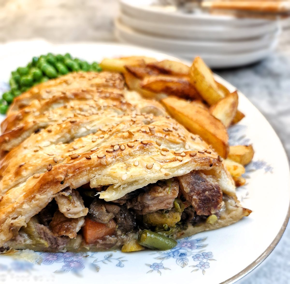 A slice of leftover roast dinner pie on a plate with chips and peas.