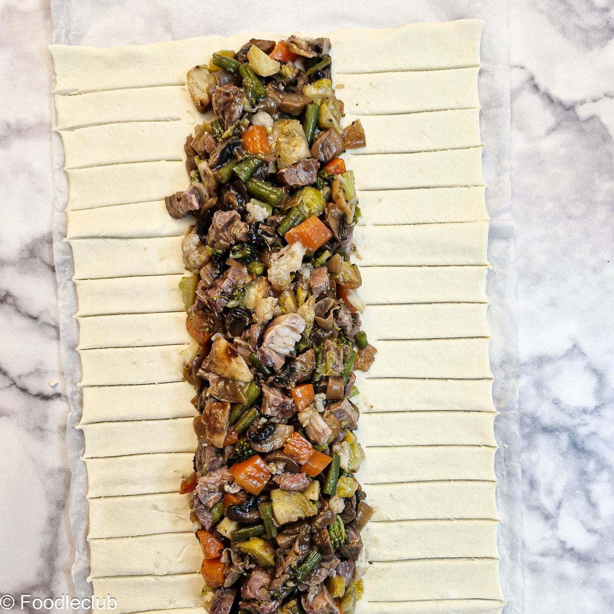 Chopped leftover meat and vegetables arranged in the centre of a sheet of puff pastry with strips cut into the pastry.