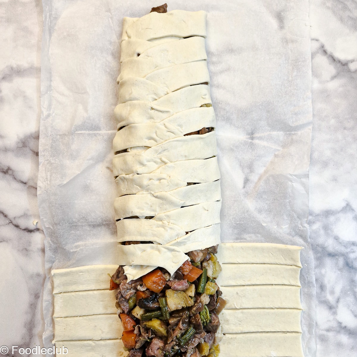 A half-assembled puff pastry braid showing how the strips of puff pastry are folded over the meat and vegetable filling.