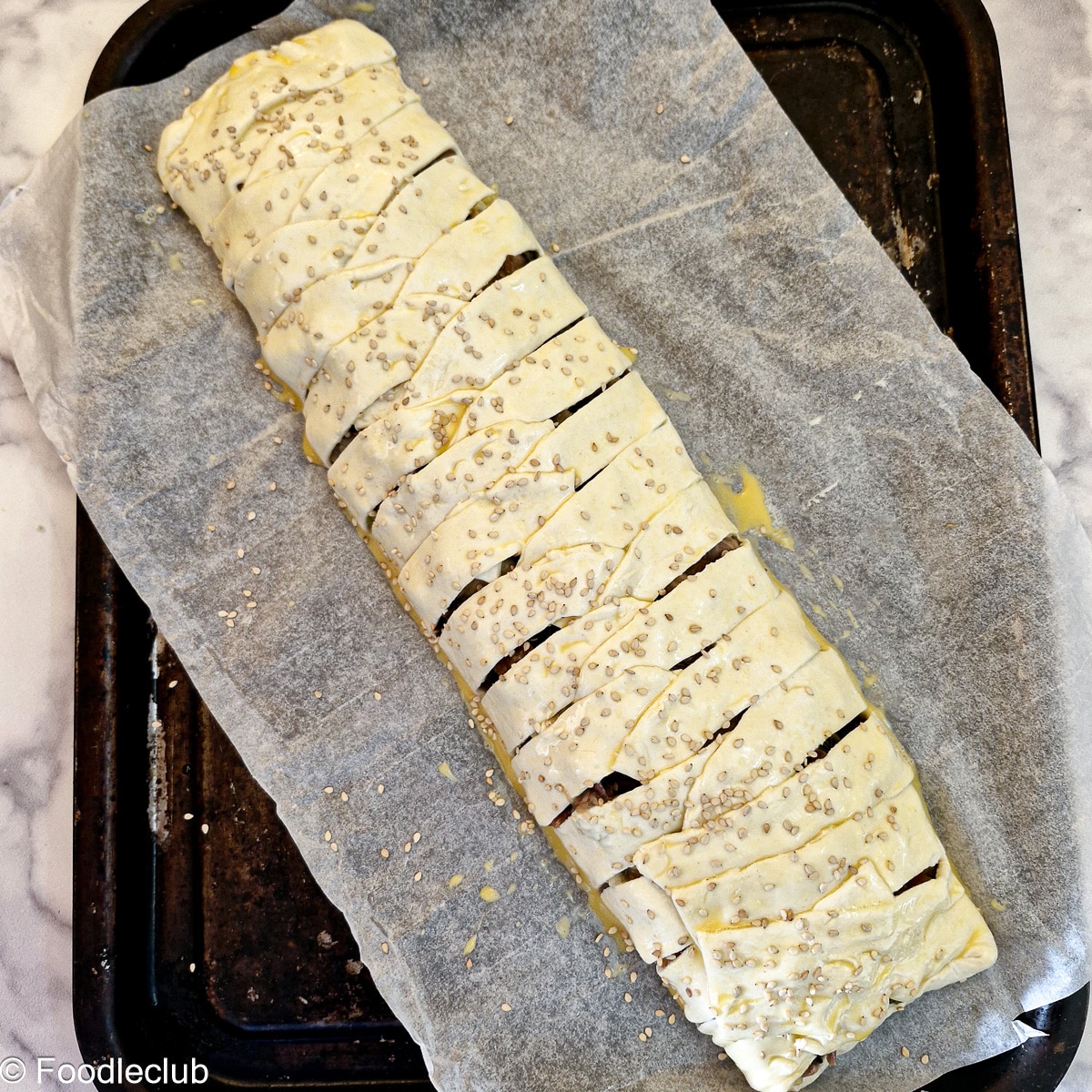 A puff pastry pie on brushed with beaten egg and sprinkled with sesame seeds on a baking tray.