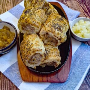 A wooden serving plate piled with stilton, pear and walnut sausage rolls alongside dishes of chutney and pickled onions.