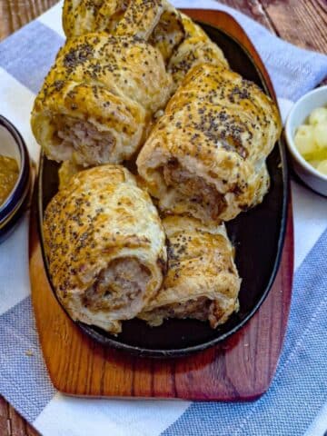 A wooden serving plate piled with stilton, pear and walnut sausage rolls alongside dishes of chutney and pickled onions.