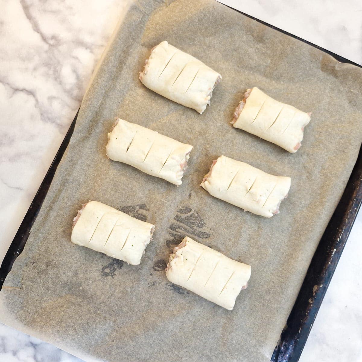 6 sausage rolls on a baking tray, with slits cut into the top of each one.