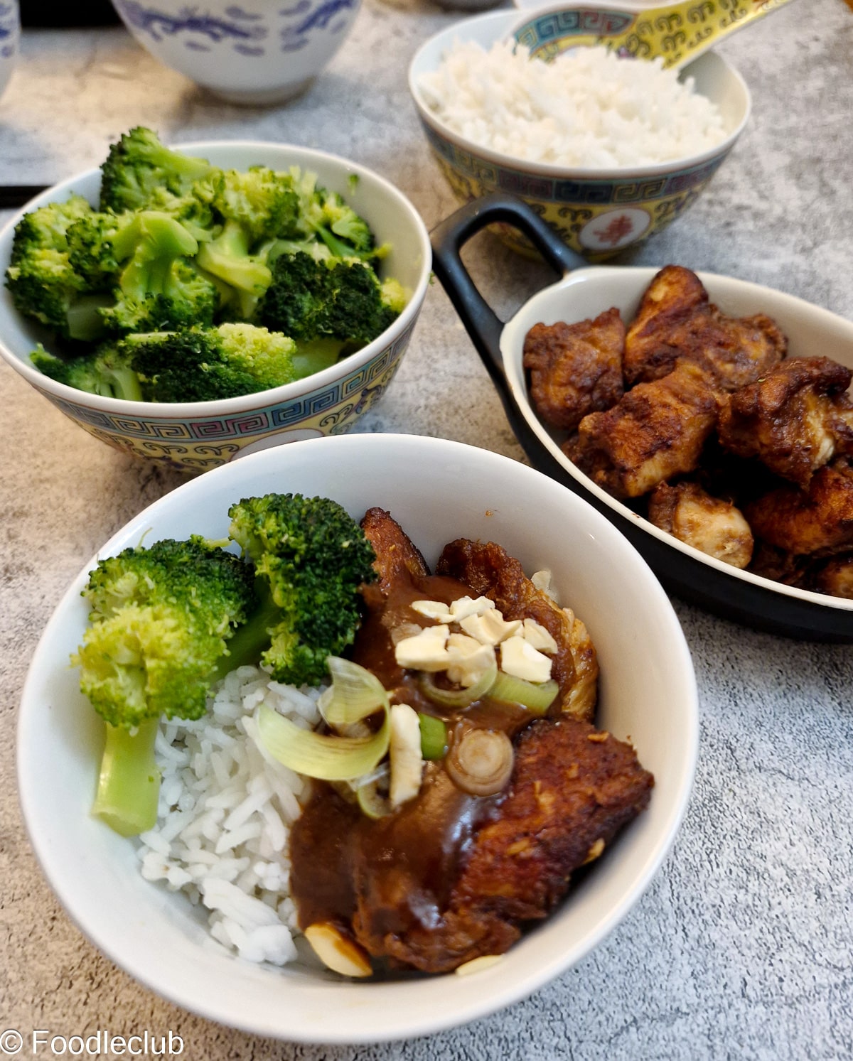 A small white bowl containing rice, broccoli and chicken covered in satay sauce. There are dishes of rice, broccoli and chicken in the background.