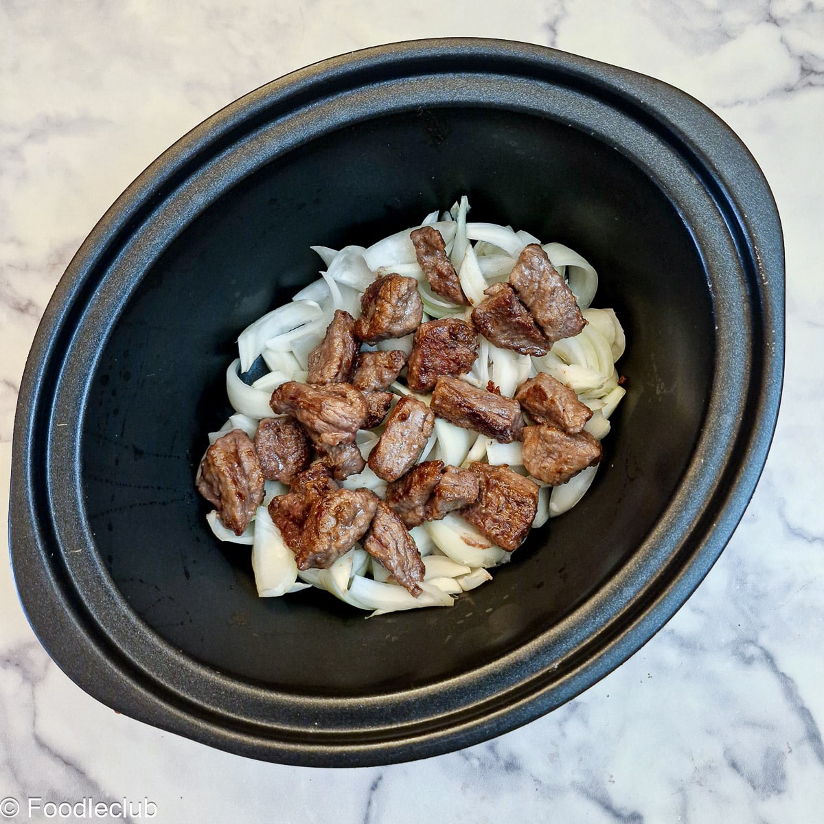 Chopped onions topped with steak pieces in the inner liner of a slow cooker.