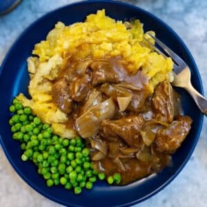 A blue dinner plate with peas and a helping of mashed potatoes smothered with braised steak and onions.