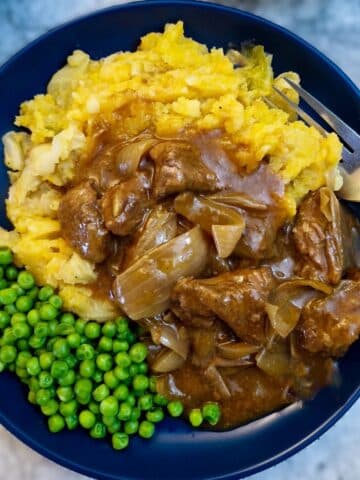 A blue dinner plate with peas and a helping of mashed potatoes smothered with braised steak and onions.