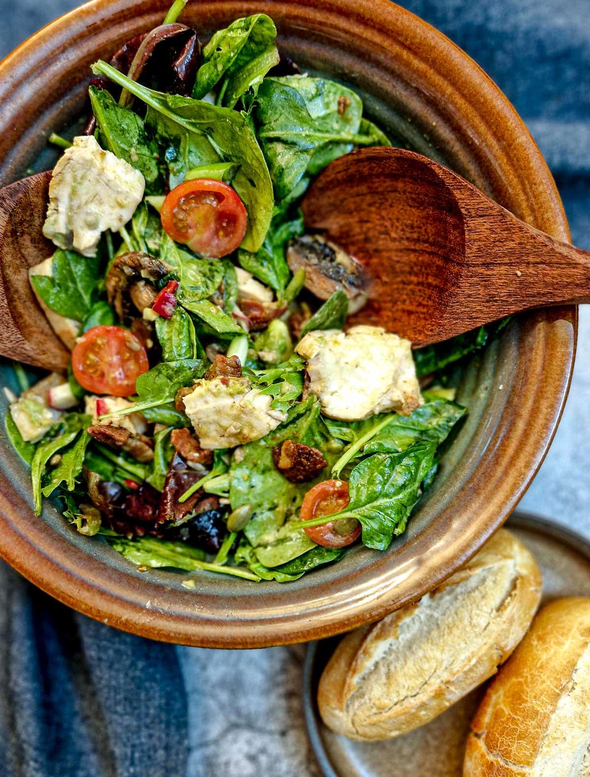 Closeup of a brown serving bowl of chicken and bacon salad. There are 2 crusty bread rolls in the foreground.