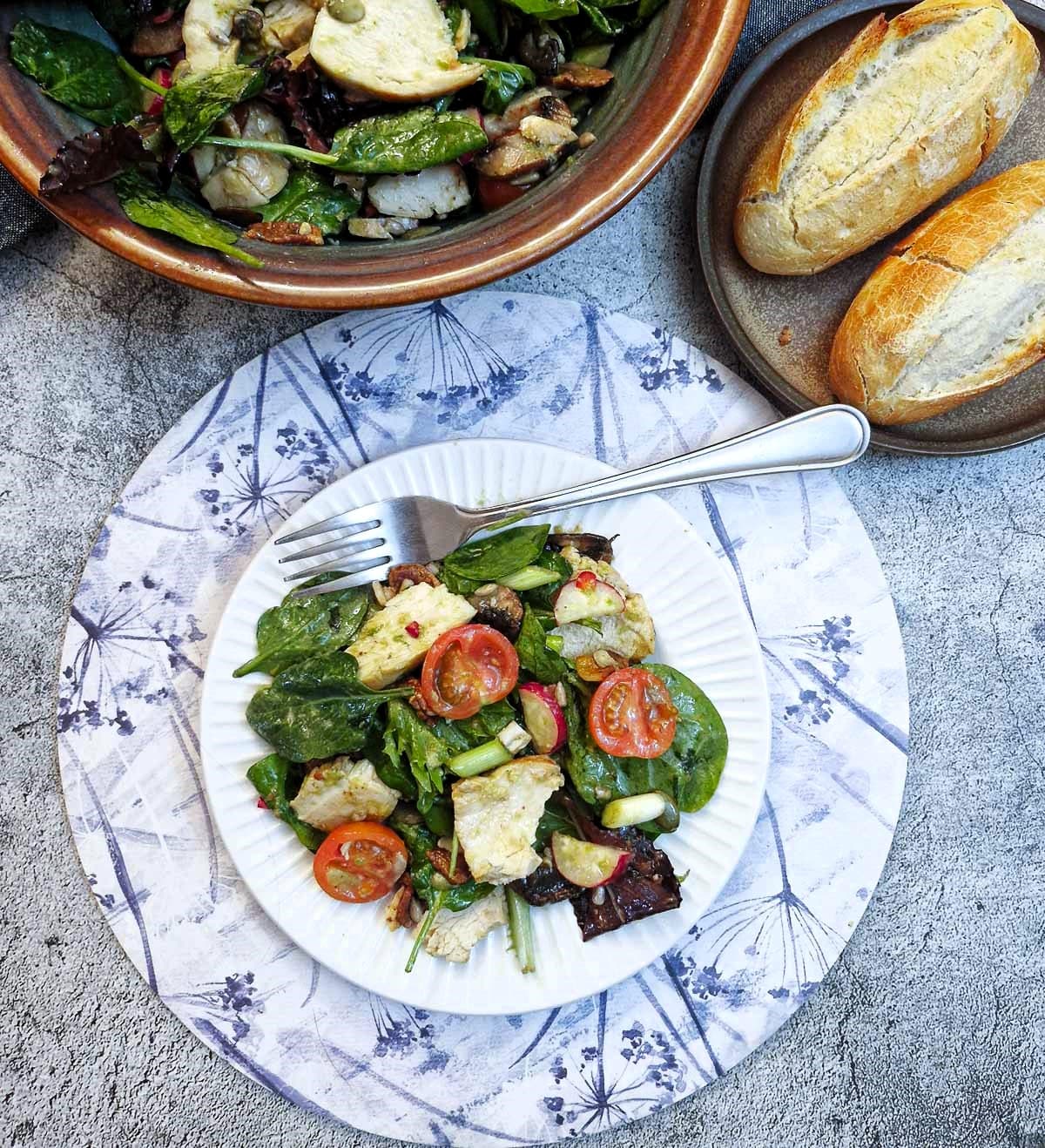 Chicken and bacon salad on a white plate. There is a bowl of salad and crusty bread in the background.