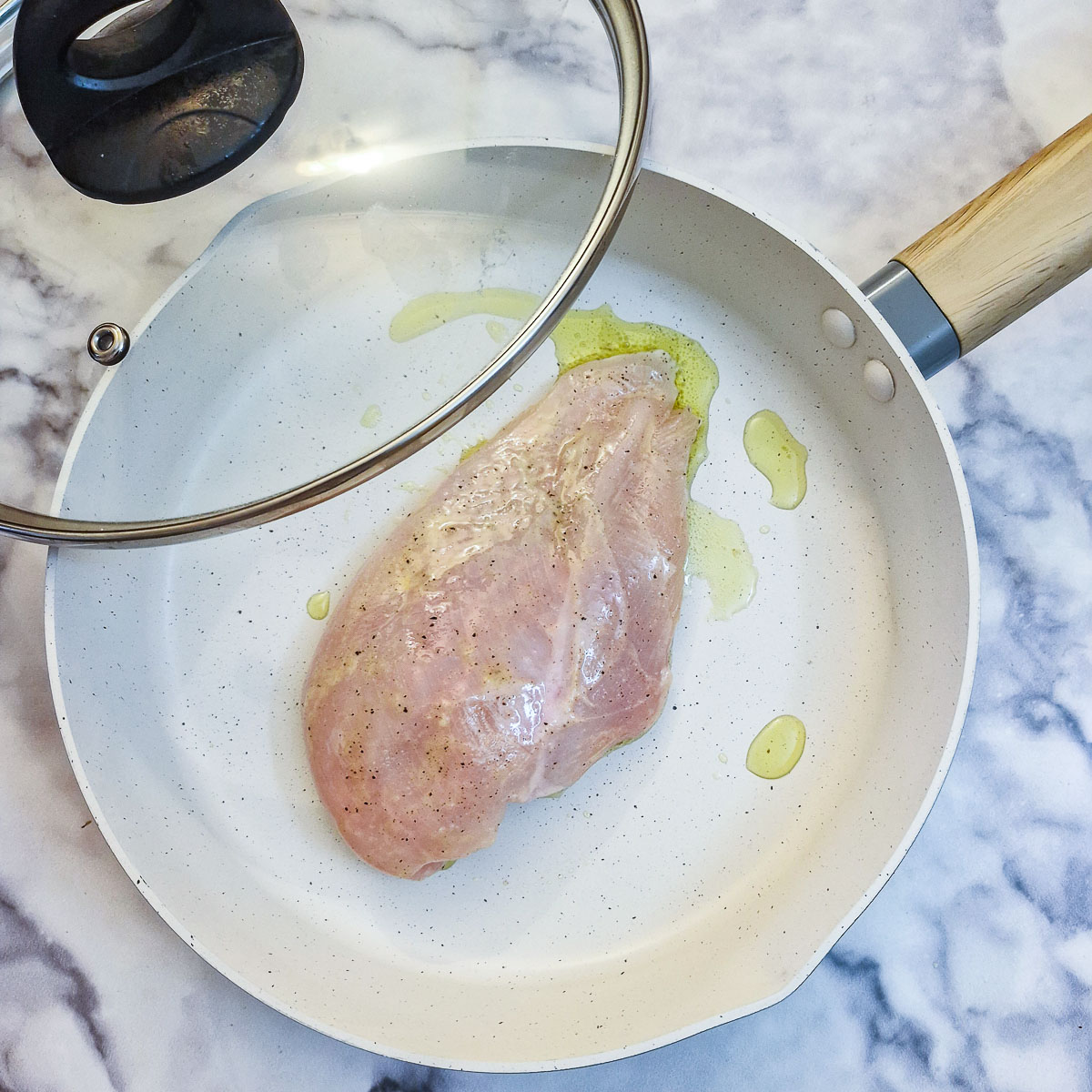 A raw chicken breast in a white frying pan with the lid partly over the pan.