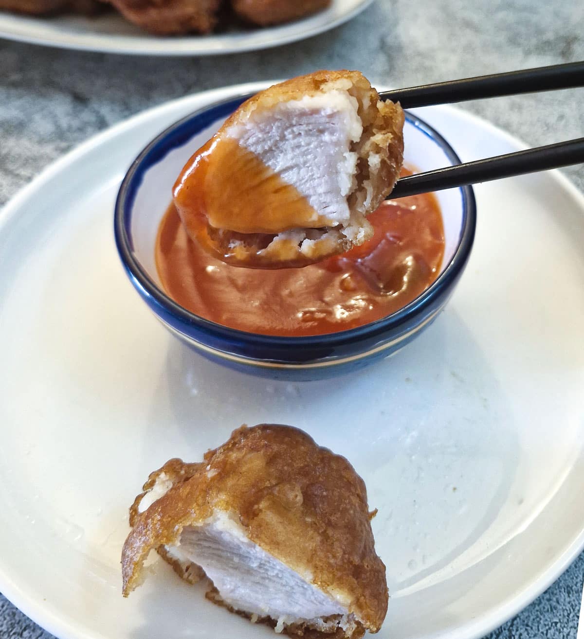 Half a crispy chicken ball being dipped into sweet and sour sauce with chopsticks.