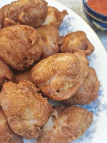 A pile of crispy chicken balls on a serving plate. There is a bowl of sweet and sour sauce in the upper right-hand corner.