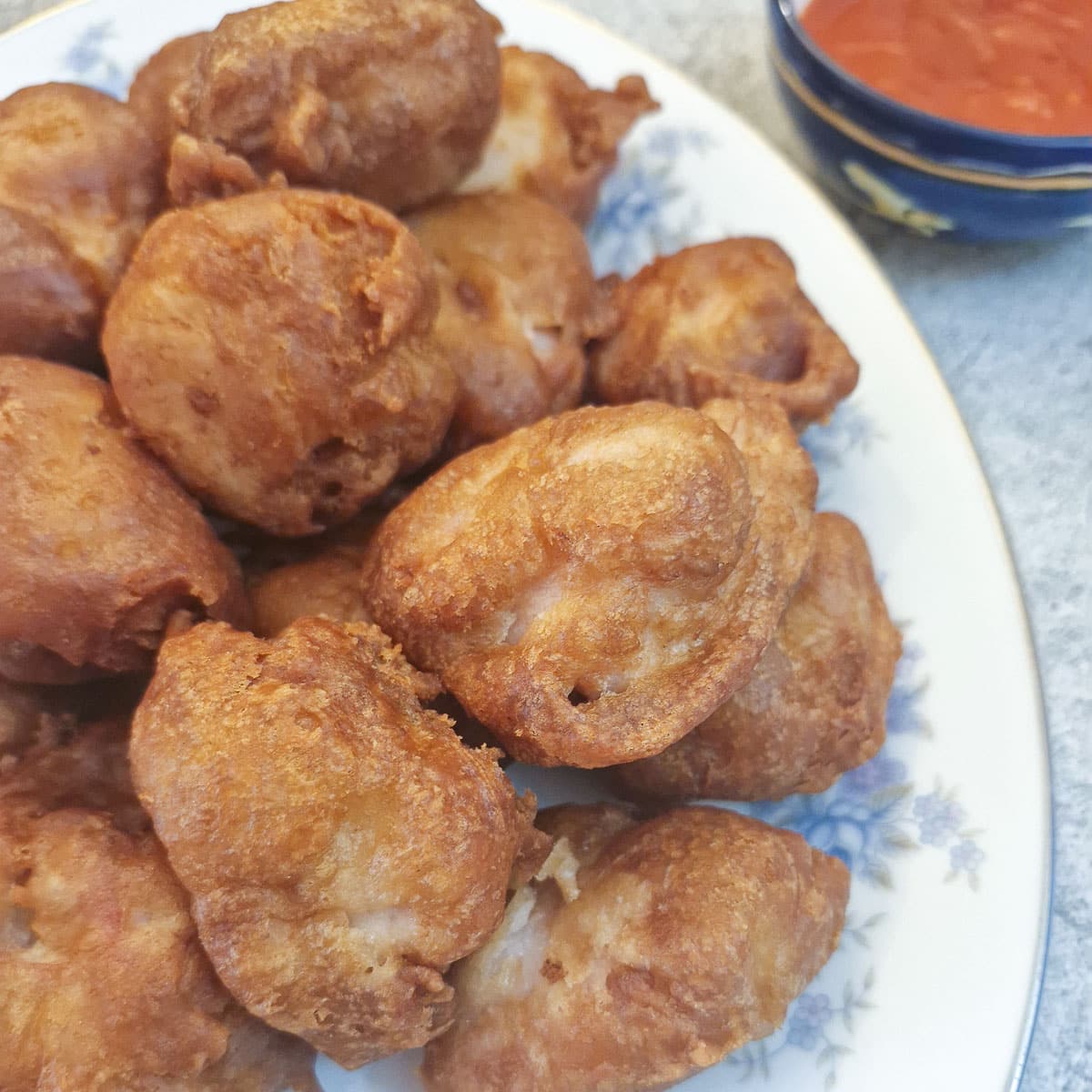 A pile of crispy chicken balls on a serving plate. There is a bowl of sweet and sour sauce in the upper right-hand corner.