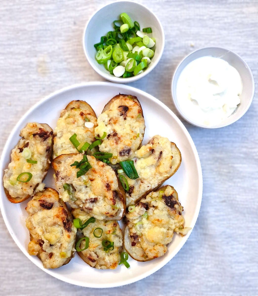 Stuffed potato skins on a white plate with a dish of sour cream and a dish of chopped spring onions.