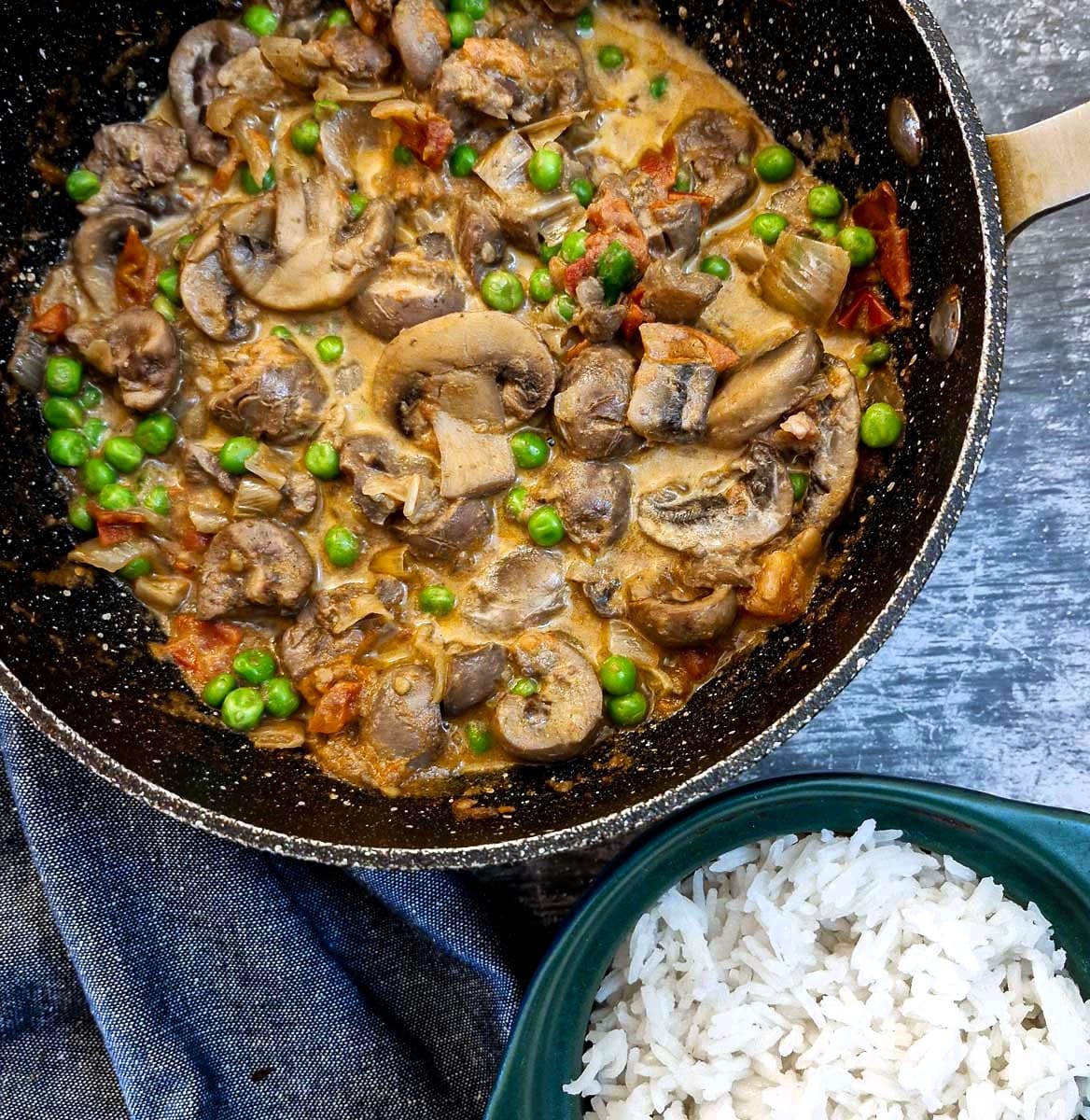 A frying pan filled with braised lamb kidneys and mushrooms with a few peas for colour.  There is a bowl of white rice in the foreground.