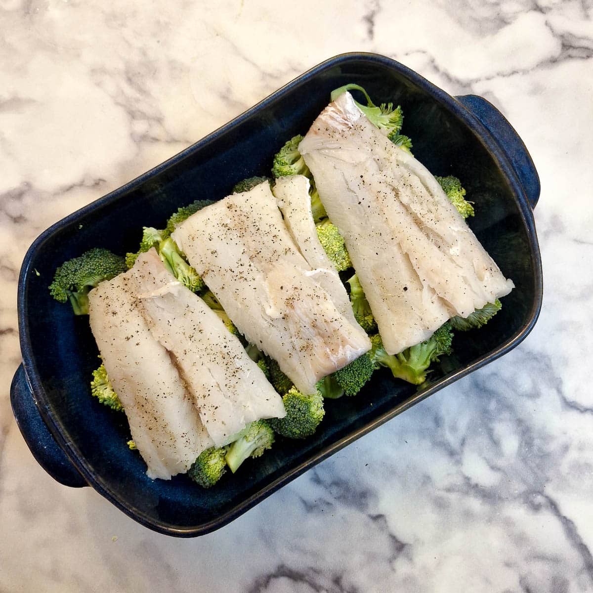 3 cod fillets on a bed of broccoli florets in a blue casserole dish.