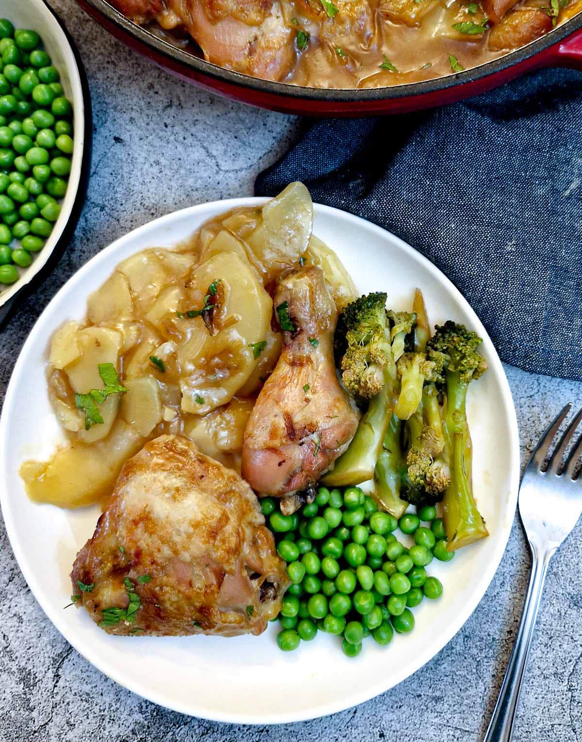 Two pieces of chicken on a white plate with french onion cooked potatoes, peas and broccoli.