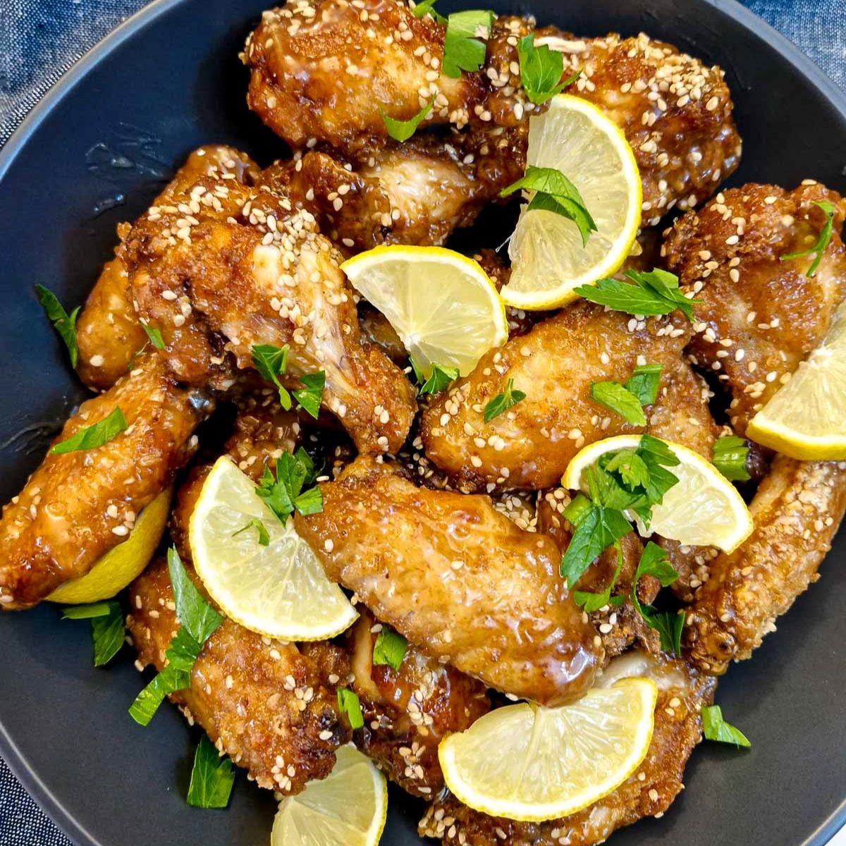 A black serving dish piled with honey lemon chiicken wings garnished with lemon slices and chopped parsley.