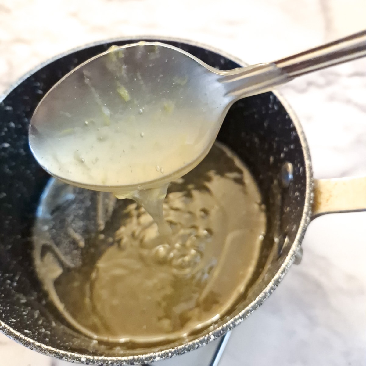 A spoon held over a pan of honey lemon sauce showing the consistency.