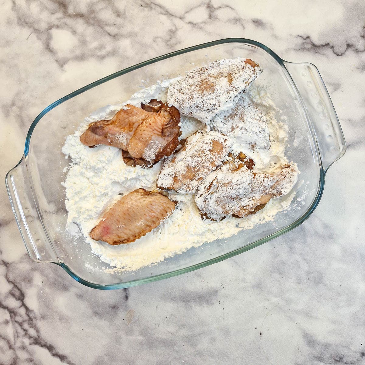 Chicken wings being coated with cornflour in a glass dish.