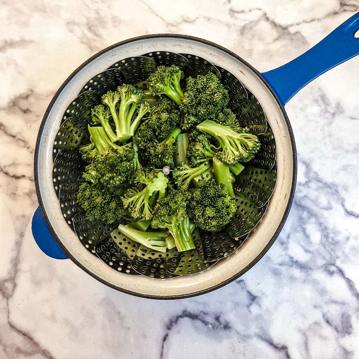 Broccoli florets in a steaming basket in a blue saucepan.