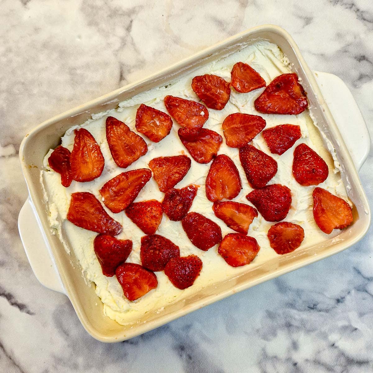 A layer of cream filling covered with sliced strawberries covering a layer of biscuits in a dish.