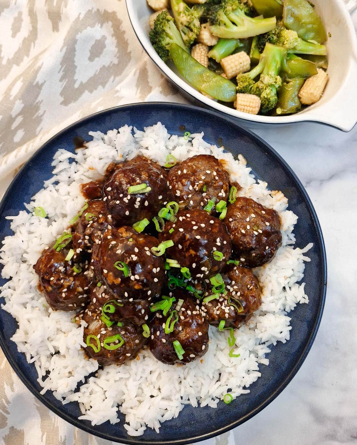Meatballs on a bed of rice in a serving dish with oyster sauce steamed vegetables in the backgrouind.