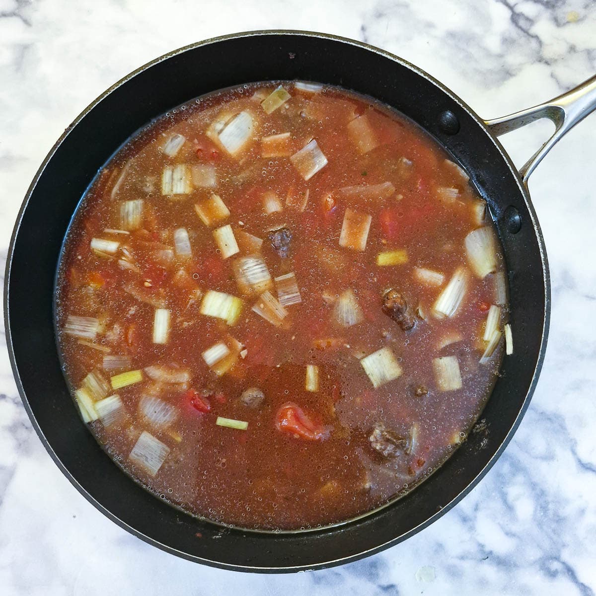 Stock, tomatoes and spices added to meat and onions in a frying pan.