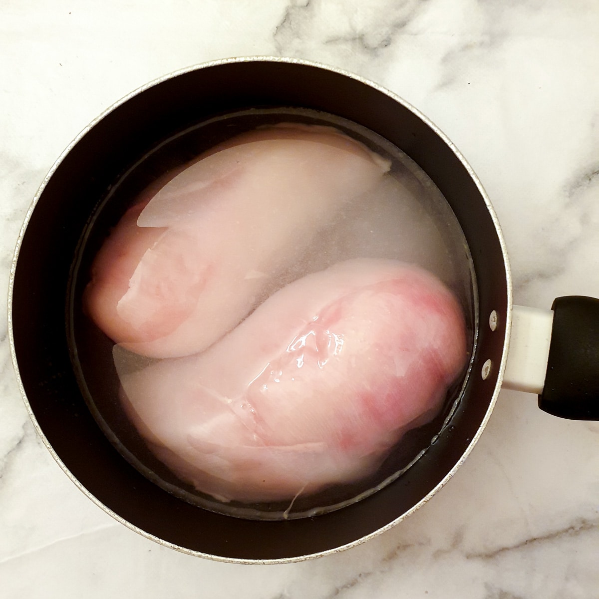 Two chicken breasts in a saucepan of water.