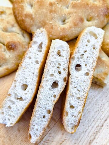 Three slices of a focaccia bread roll in front of a pile of uncut rolls.