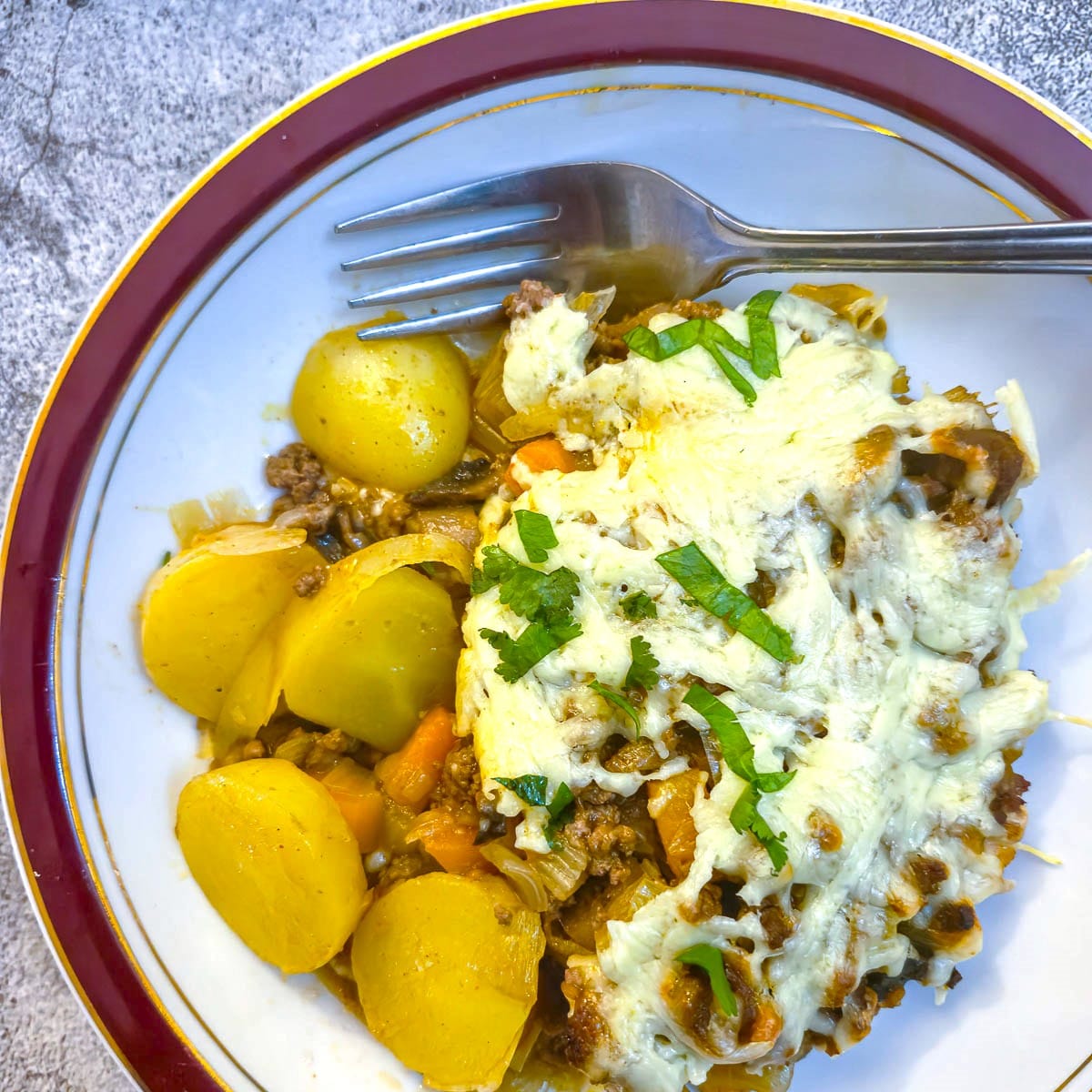 Close up of mince and potato bake in a red-rimmed dish, with sliced potatoes on the side.