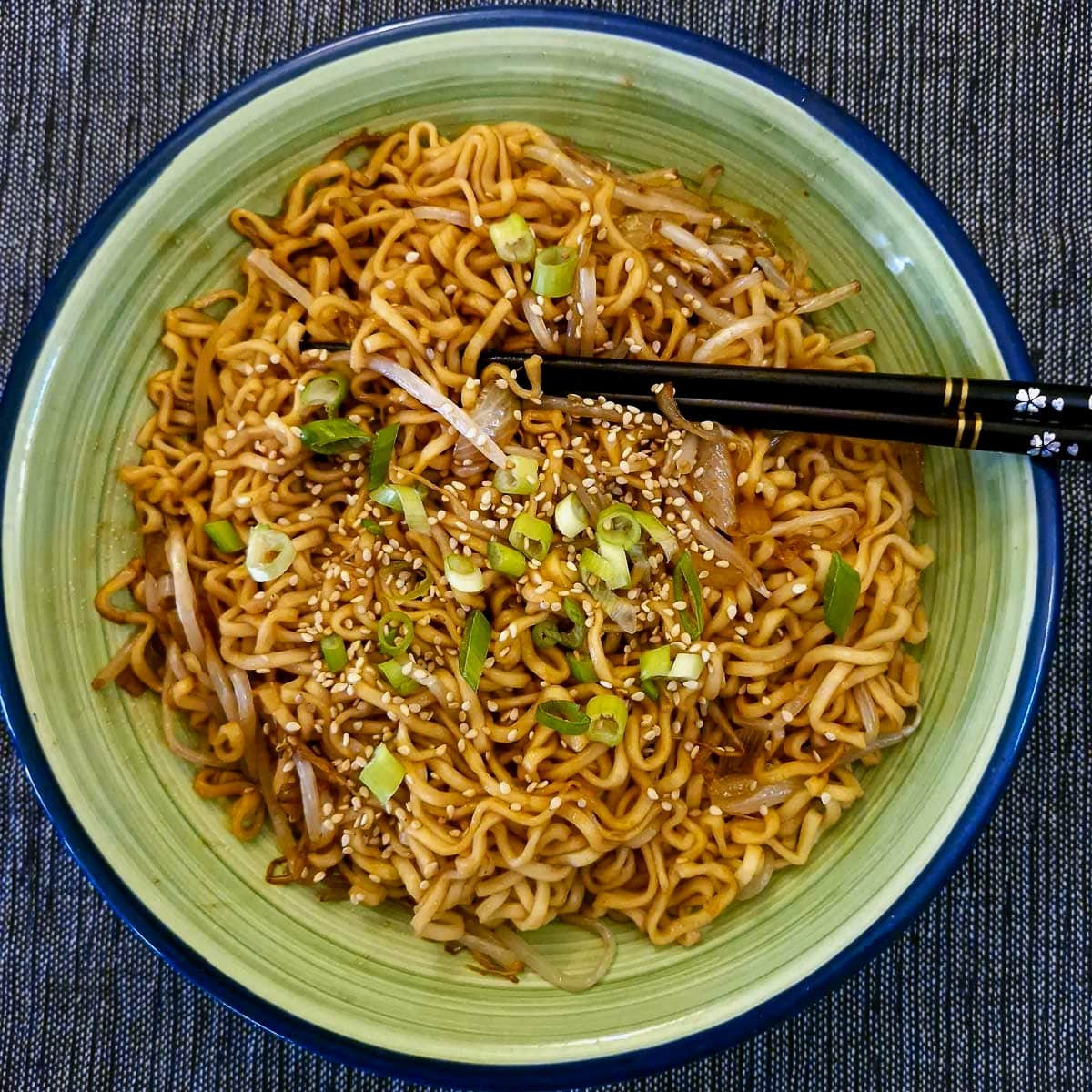 A green serving dish of Chinese noodles with a pair of black chopsticks.