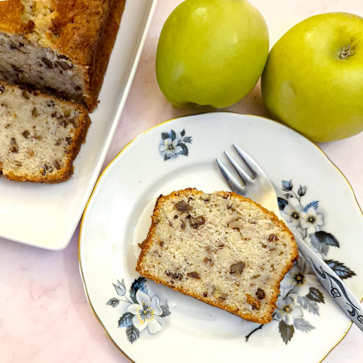 A slice of apple and pecan nut loaf on a blue patterned plate. There are 2 apples in the background.