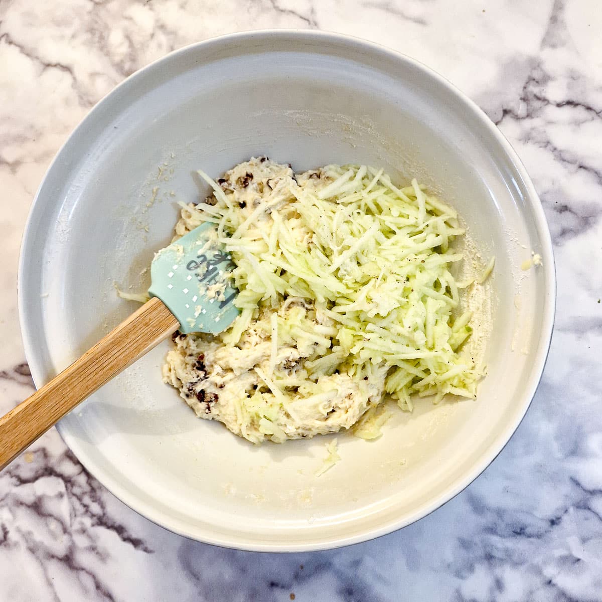 Grated apples on top of the cake batter in a white mixing bowl.