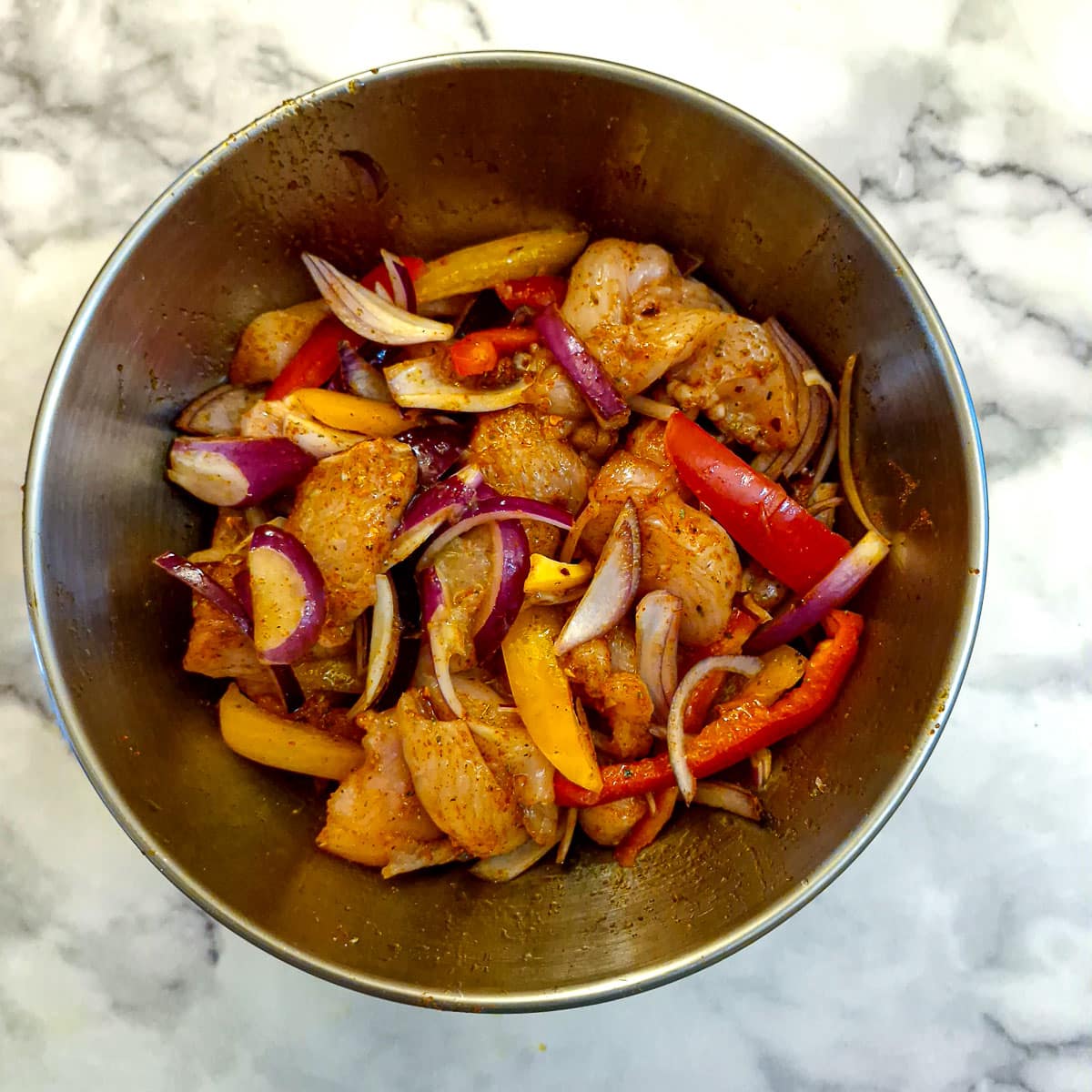 Sliced chicken, capsicums and onions coated with spices in a stainless steel mixing bowl.