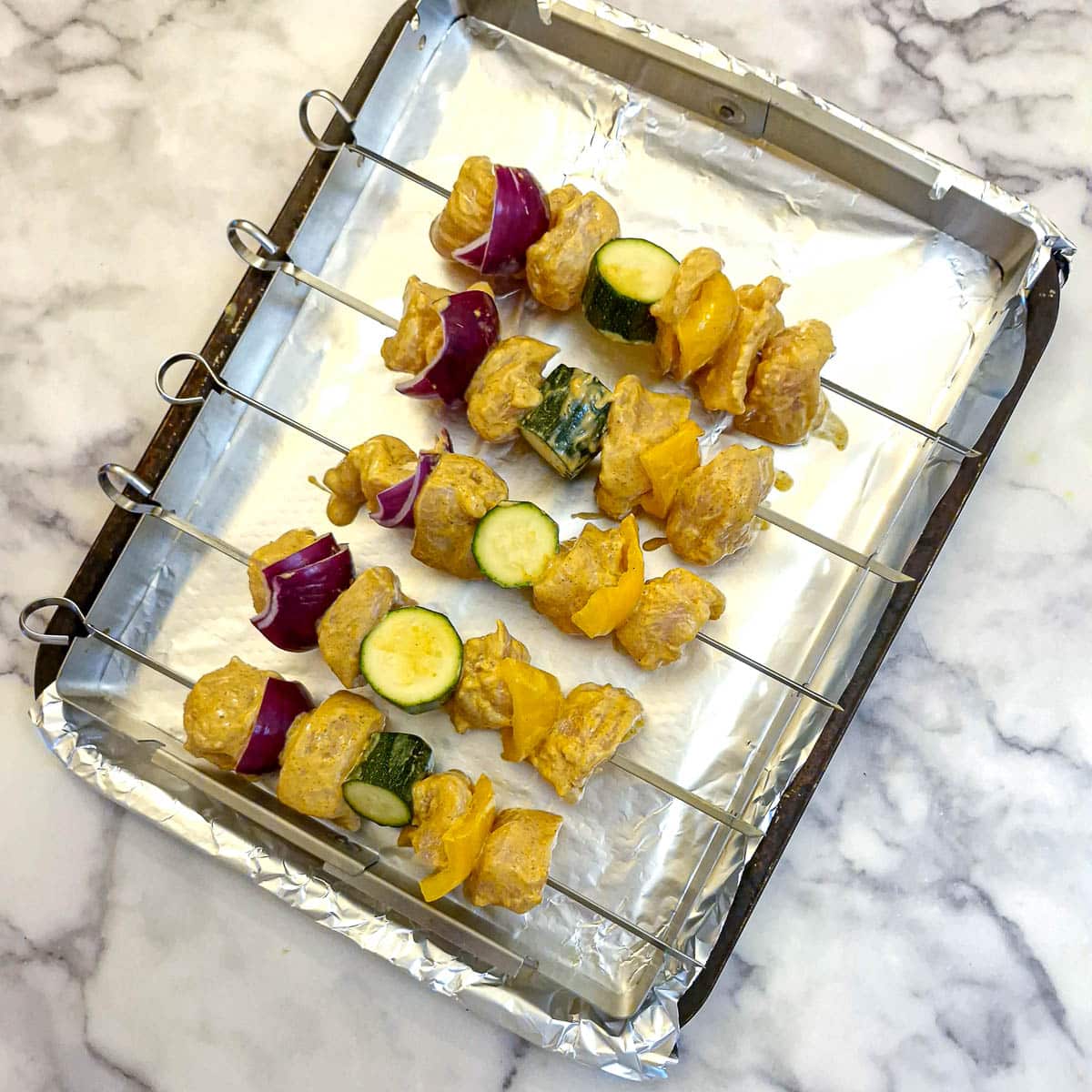 5 uncooked chicken skewers arranged on a rack set on a baking tray.