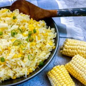 A dish of buttered corn and rice with a wooden serving spoon. There are 2 corn cobs in the foreground.