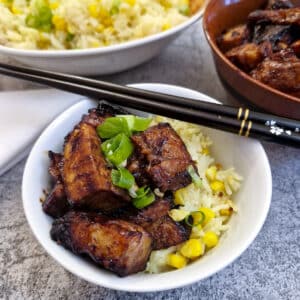 A white bowl of pork belly bites, with rice. There is a pair of chopsticks balanced across the bowl.