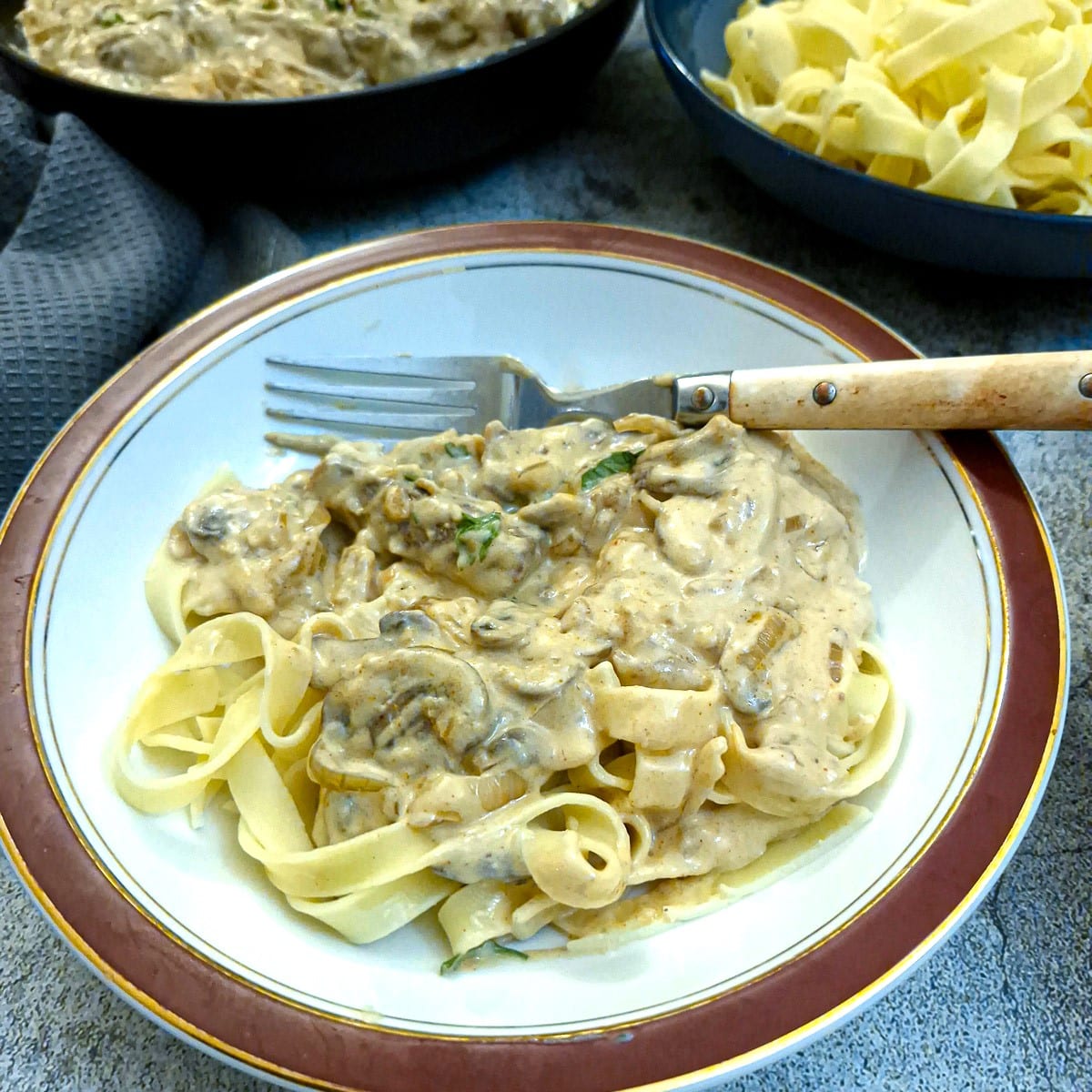 A white red-rimmed bowl filled with beef and mushroom stroganoff on a bed of tagliatelle.